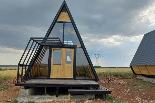 Teraslı ve pergoleli üçgen bungalov bir tarla üzerinde duruyor, büyük camlarıyla bu üçgen bungalovun doğayla uyumunu izliyoruz.